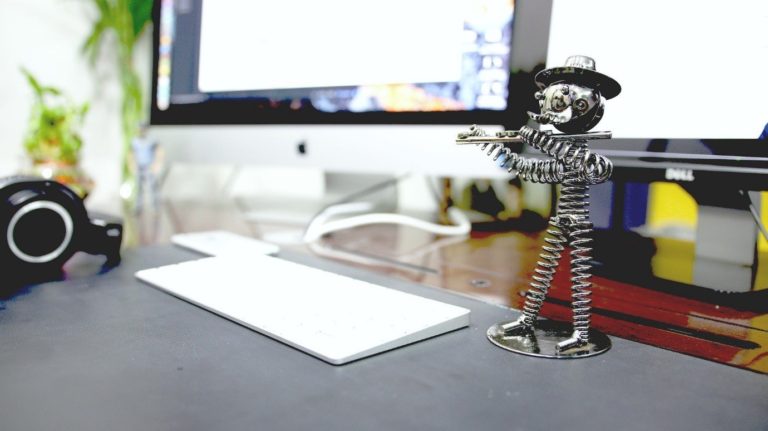 Foto scrivania grigia con tastiera pc bianca e statuina in acciaio inox di un omino col cappello che suona il flauto. Piante verdi sullo sfondo.